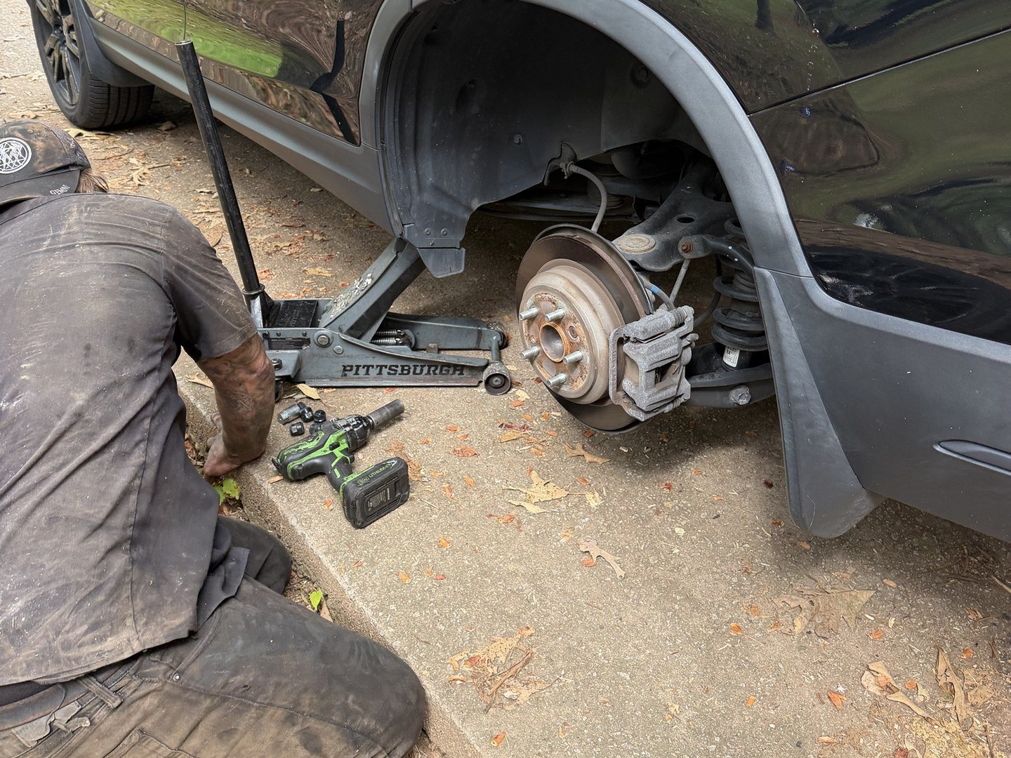 Mechanic kneeling at the curb beside an SUV on a jack with the rear wheel off and the brake rotor exposed